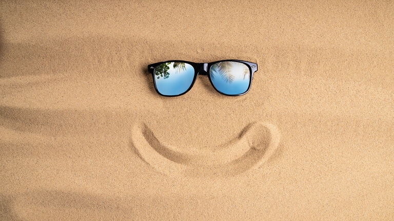 Sonnenbrille im Sand, darunter ein in den Sand gemalter lächelnder Mund, sodass ein Smiley entsteht