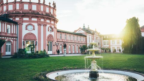 Das Schloss Biebrich von außen bei niedrigstehender Sonne mit einem Springbrunnen am vorderen Bildrand