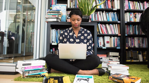 Eine junge PoC-Frau sitzt mit einem Laptop auf dem Schoß vor einem Bücherregal, umgeben von Büchern