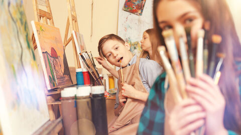 Der Wettbewerb „Jugend malt“ soll die kreative Entwicklung junger Menschen fördern Ein Mädchen hält Pinsel in die Kamera, hinter ihr schaut ein Kindhervor, beide sitzen vor Leinwänden