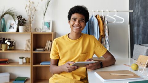 "Ohne Punkt und Komma" soll die Schreiblust fördern PoC-Jugendlicher sitzt am Schreibtisch mit Tablet und Schreibblock, lächelt zugewandt in die Kamera
