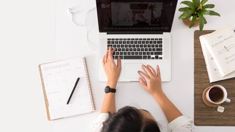 Sprechen Sie uns an! Schreibtisch aus der Vogelperspektive. Eine Frau sitzt am Laptop, rechts steht eine Pflanze, eine Tasse Tee, ein Block liegt bereit