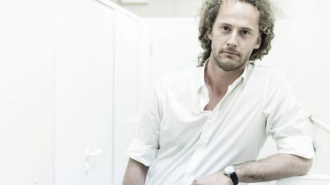 Porträt von Florian Lutz. Er trägt ein weißes Hemd vor weißem Hintergrund und hat seinen Arm aufgestützt