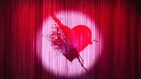 Spotlight auf einen roten Vorhang, im Scheinwerferlicht ist ein Herz zu sehen, durch den ein Pfeil geht