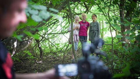 Das mobile Filmstudio und seine Initiatorinnen und Initiatoren unterstützen die Teilnehmenden dabei, gemeinsam Idee und Drehbuch zu entwickeln und dann technisch umzusetzen. Zwei Mädchen im Wald, im Vordergrund steht eine Kamera