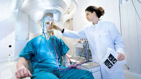Ein Mann im blauen Hygieneanzug trägt eine Kappe, die Sensoren an seinem Kopf hält. Er sitzt unter einer Maschine mit einer Aussparung für seinen Kopf. Neben ihm steht eine Frau im weißen Kittel, die seine Maske justiert