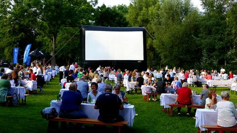 Gäste sitzen an Tischen auf einer Wiese, im Hintergrund ist eine Kinoleinwand aufgebaut