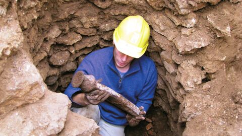 Blick in ein Erdloch; dort steht ein Mann mit gelbem Schutzhelm, er hält ein römisches Bleirohr in den Händen