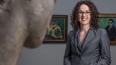 Angela Dorn im Museum, lächelnd, im Vordergrund ist eine Statue angeschnitten, im Hintergrund hängen Bilder