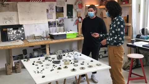 Ministerin Dorn steht mit einem Künstler an einem Tisch, auf dem verschiedene Kunstobjekte aus Mikroplastik drapiert sind