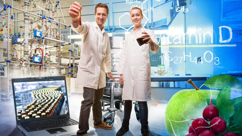 Composing: Ein Mann und eine Frau in Laborkitteln halten Reagenzgläser in die Kamera, drum herum sind chemische Formeln und Laboraccessoires drapiert