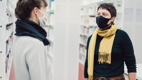 Im Wintersemester 2021/2022 soll an Hessens Hochschulen die Präsenzlehre wieder möglichst zum Regelfall werden.  Zwei Frauen, beide mit Masken, stehen in einer Universitätsbibliothek und lächeln sich an