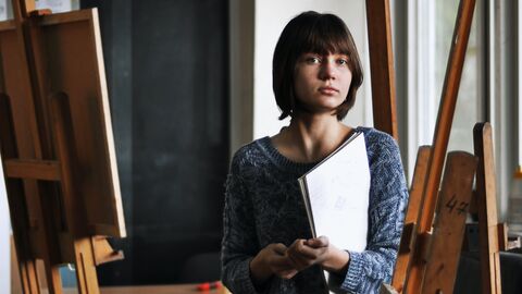 Professionelle Künstlerinnen sind in der Wahrnehmung der Öffentlichkeit immer noch zu wenig präsent - das wollen wir ändern Eine Künstlerin in ihrem Atelier, sie hält einen Zeichenblock in der Hand und schaut ernst