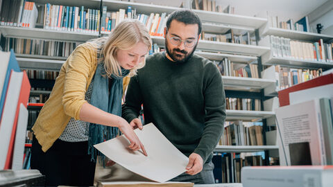 Kontakte knüpfen und Neues lernen Zwei Studierende stehen vor einem Bücherregal und betrachten einen historischen Bogen Papier