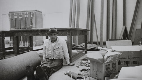 Schwarzweiß-Foto von Joseph Beuys, er sitzt, umgeben von Arbeitsmaterialien, auf dem Boden eines Ausstellungsraums