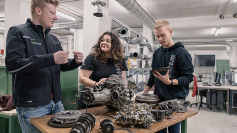 Drei Studierende stehen um einen Tisch herum, auf dem Einzelteile eines Rennautos liegen