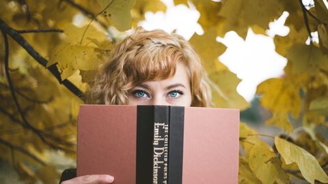 Eine Frau hält sich ein Buch vors Gesicht, im HIntergrund sieht man Bäume und Sonnenschein