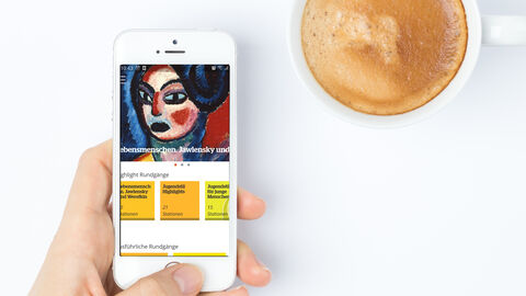 Die hessischen Landesmuseen, Schlösser und Gärten haben ein großes digitales Angebot  Eine Hand mit Smartphone, auf dem das Onlineangebot des Museums Wiesbaden zu sehen ist