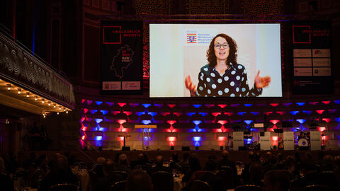 Der Festsaal im Wiesbadener Kurhaus mit barockem Schmuck, festlichbeleuchtet; eine Leinwand ist von der Decke gelassen, auf der ein Grußwort-Video von Ministerin Dorn abgespielt wird 