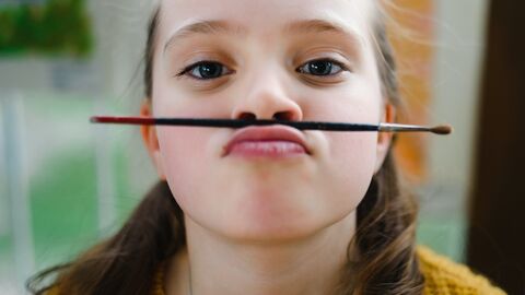 Ein Mädchen hat sich einen Pinsel zwischen Nase und Oberlippe geklemmt und grinst in die Kamera