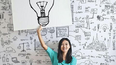 Hessen Ideen weckt den Gründergeist an den Hochschulen! Eine Frau sitzt vor einer Graffitiwand und hält ein Schild in die Höhe, auf dem eine Glühbirne zu sehen ist