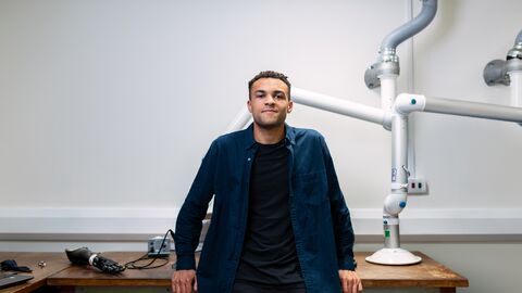 Ein junger Mann steht an einem Arbeitstisch und lächelt selbstbewusst in die Kamera