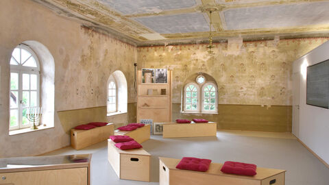 Innenraum der ehemaligen Synagoge in  Meißner-Abterode: An den Wänden sind goldene Ornamente freigelegt, es stehen Sitzbänke und ein Bücherregal im Raum