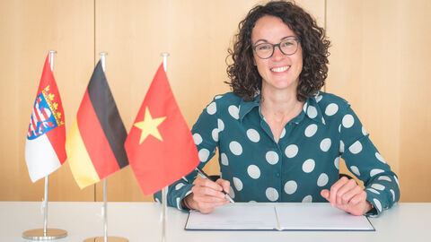 Ministerin Dorn sitzt am Schreibtisch, vor ihr die Fahnen von Hessen, Deutschland und Vietnam; sie unterzeichnet das Abkommen