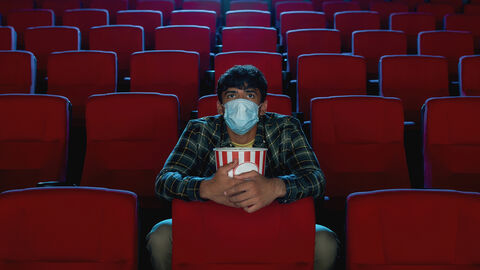 Ein Mann sitzt allein im Kinosaal, er trägt eine Maske und hat einen Eimer Popcorn vor sich
