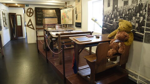 Hier wurde früher gelernt: ein original Klassenzimmer aus vergangenen Zeiten im Waldecker Spielzeugmuseum. Blick ins Waldecker Spielzeugmuseum: Man sieht ein historisches Klassenzimmer mit Bänken, Pult und zwei großen Teddybären