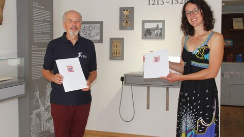 Kunst- und Kulturministerin Angela Dorn hat Joachim Hübner, Vorsitzender des Kultur- und Geschichtsvereins Bad Emstal e. V. die Urkunde und einen Bescheid über 1.000 Euro überreicht. Kunst- und Kulturministerin Angela Dorn und Joachim Hübner, Vorsitzender des Kultur- und Geschichtsvereins Bad Emstal e. V., stehen mit Plakette und Urkunde vor einer Wand mit Ausstellungsstücken im Museum.