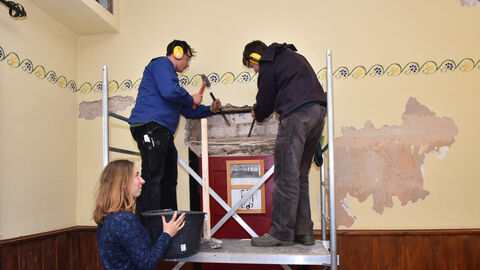 Zwei Personen stehen auf einem Gerüst und meißeln eine Wand frei, darunter liegt altes Fachwerk