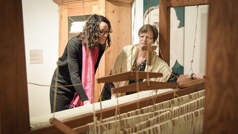 Museumsbeauftragte Elisabeth Rößler (r.) zeigt Kunst- und Kulturministerin Angela Dorn den Webstuhl.  Ministerin Dorn arbeitet an einem großen Webstuhl, die Museumsbeauftragte steht daneben
