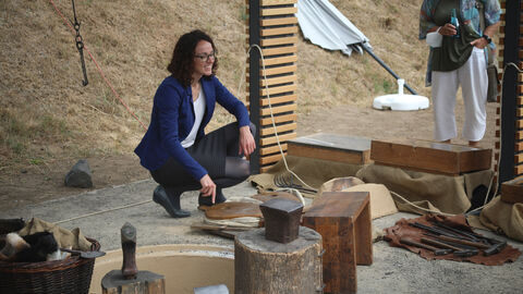 Ministerin Dorn benutzt einen Blasebalg in der keltischen Schmiede der Keltenwelt am Glauberg, um sie herum sind Amboss und Werkzeuge verteilt