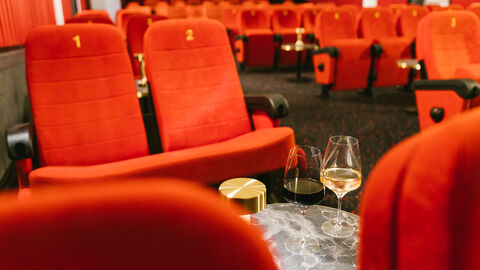 Der Hessische Film- und Kinopreis wird in diesem Jahr Ende November in drei Veranstaltungen in Kinos in Kassel, Bad Nauheim und Frankfurt vergeben. Blick in den Kinosaal der FilmBühne Bad Nauheim, man sieht rote Kinosessel mit kleinen Tischen davor, auf einem stehen zwei Weingläser
