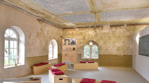 Innenraum der ehemaligen Synagoge in  Meißner-Abterode: An den Wänden sind goldene Ornamente freigelegt, es stehen Sitzbänke und ein Bücherregal im Raum