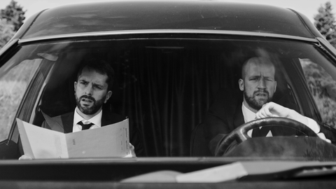 Szene aus "Nemesis" Szene aus "Nemesis": Eine Schwarzweißszene; zwei Männer sitzen in einem Auto, einer liest in einer Mappe