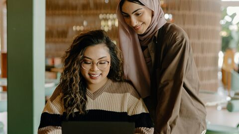 Eine Frau mit dunklen Locken und eine Frau mit Kopftuch schauen in einen Laptop