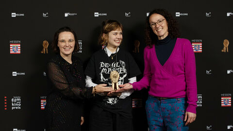 Filmchefin Anna Schoeppe und Ministerin Angela Dorn stehen mit Preisträgerin Lea Drinda vor einer Fotoleinwand.