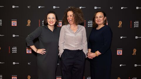 Anna Schöppe, Chefin von HessenFilm, die Regisseurin Carolin Schmitz und Staatssekretärin Ayse Asar stehen vor einer Logoleinwand.