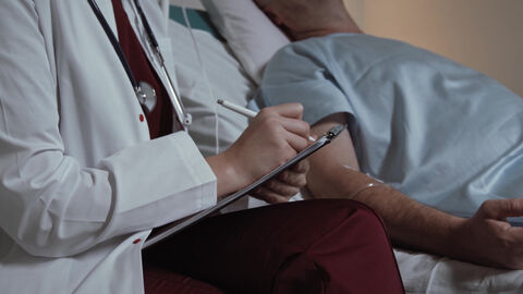 In der Klimasprechstunde geht es um Auswirkungen der Klimakrise auf die Gesundheit und darum, wie Medizinerinnen und Mediziner für ihren ökologischen Fußabdruck im Beruf sensibilisiert werden können Eine Person im weißen Kittel sitzt neben einer liegenden Person.