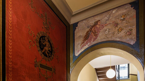 Unter der Nachkriegs-Fliesenverkleidung und Tapeten war eine bauzeittypische Dekorationsmalerei versteckt, wie sie nur selten in diesem Zustand in Frankfurt erhalten ist. Blick ins Treppenhaus: Über einem Bogendurchgang ist ein Blüten streuender Engel auf die Wand gemalt