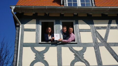 Ministerin Dorn schaut aus einem Fenster im obersten Stock des prämierten Fachwerkhauses, die Eigentümer aus dem Fenster daneben. Sie halten gemeinsam die Ehrenurkunde.