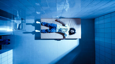 Szene aus "Zelle 5": Ein dunkelhäutiger Mann liegt angekettet auf einer Matratze in einer Gefängniszelle