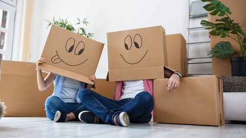 Zwei Personen sitzen auf dem Boden, umgeben von Umzugskartons. Sie tragen jeweils einen Karton als Maske auf dem Kopf, darauf sind Gesichter gemalt