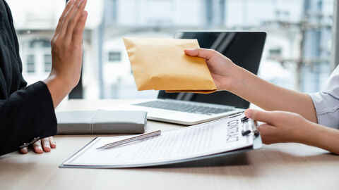 Eine Person bietet einer anderen Person einen mit Geld gefüllten Umschlag, die andere Person hebt abwährend die Hand