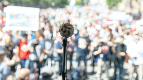 Ein Mikrofon, das auf den Betrachter gerichtet ist; im Hintergrund protestierende Menschen