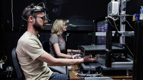 Zwei Personen in einer Laborsituation: Der Probant trägt eine Konstruktion auf dem Kopf, die ihn mit dem PC vor ihm verbindet, die Forscherin sitzt neben ihm.