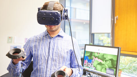 Den digitale Zwilling einer Zelle zu erstellen ist das Ziel der Clusterinitiative SCALE der Goethe-Universität Frankfurt. SCALE geht jetzt in die zweite Runde des Exzellenzstrategie-Wettbewerbs. Ein Mann mit VR-Brille und Bildschirm in Hintergrund.