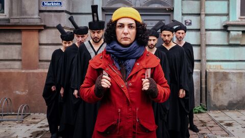 Eine Frau im roten Mantel steht vor einer Gruppe Menschen in schwarzen Togen.